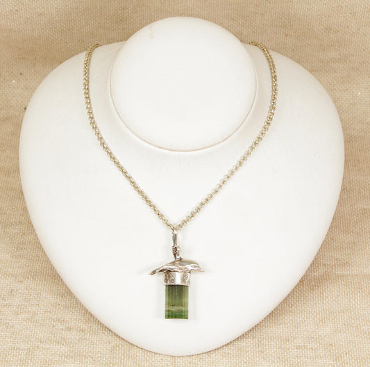 Silver dolphin necklace with a green pendant on a white heart-shaped stand against a beige background