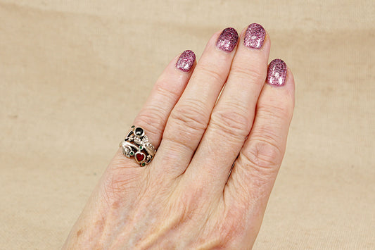 Hand with pink glittery nails wearing an art nouveau silver ring with red and black gemstones on a beige background