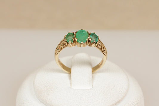 Gold ring with three green emerald gemstones on a white stand against a beige background