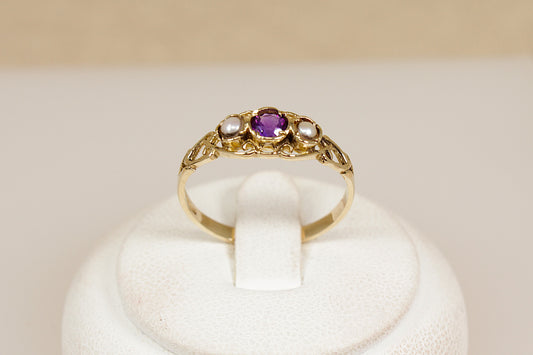 Gold ring with a purple amethyst gemstone and seed pearls on a white stand against a beige background