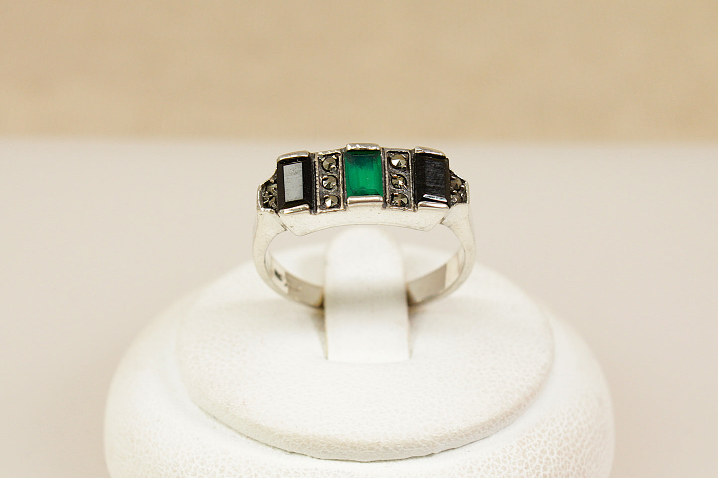 Silver art deco ring with green gemstone on a white stand against a beige background