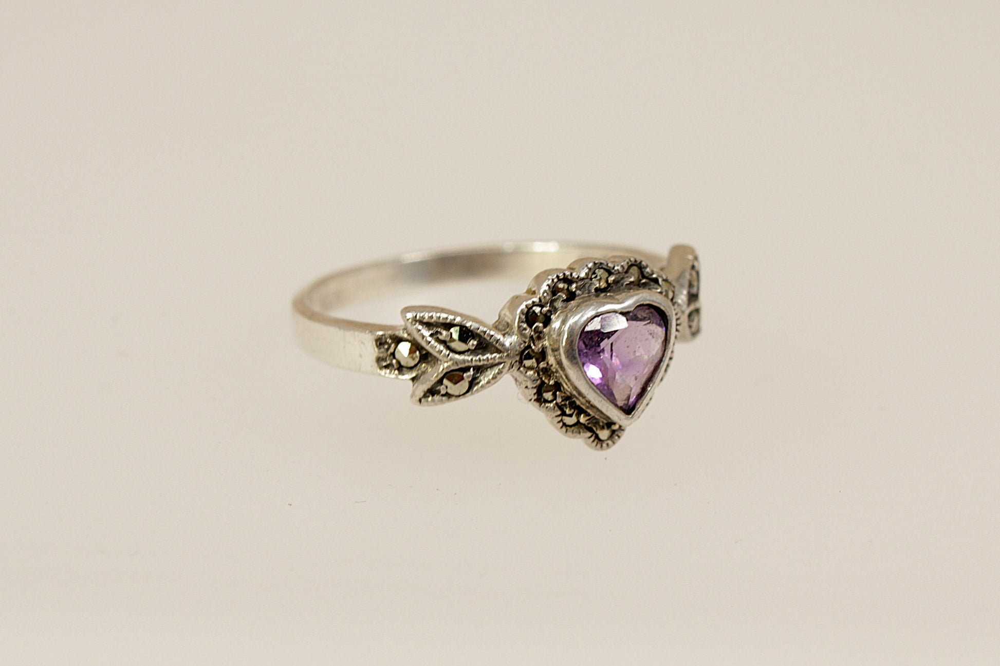 Silver ring with a heart-shaped purple amethyst and marcasite gemstones on a beige background