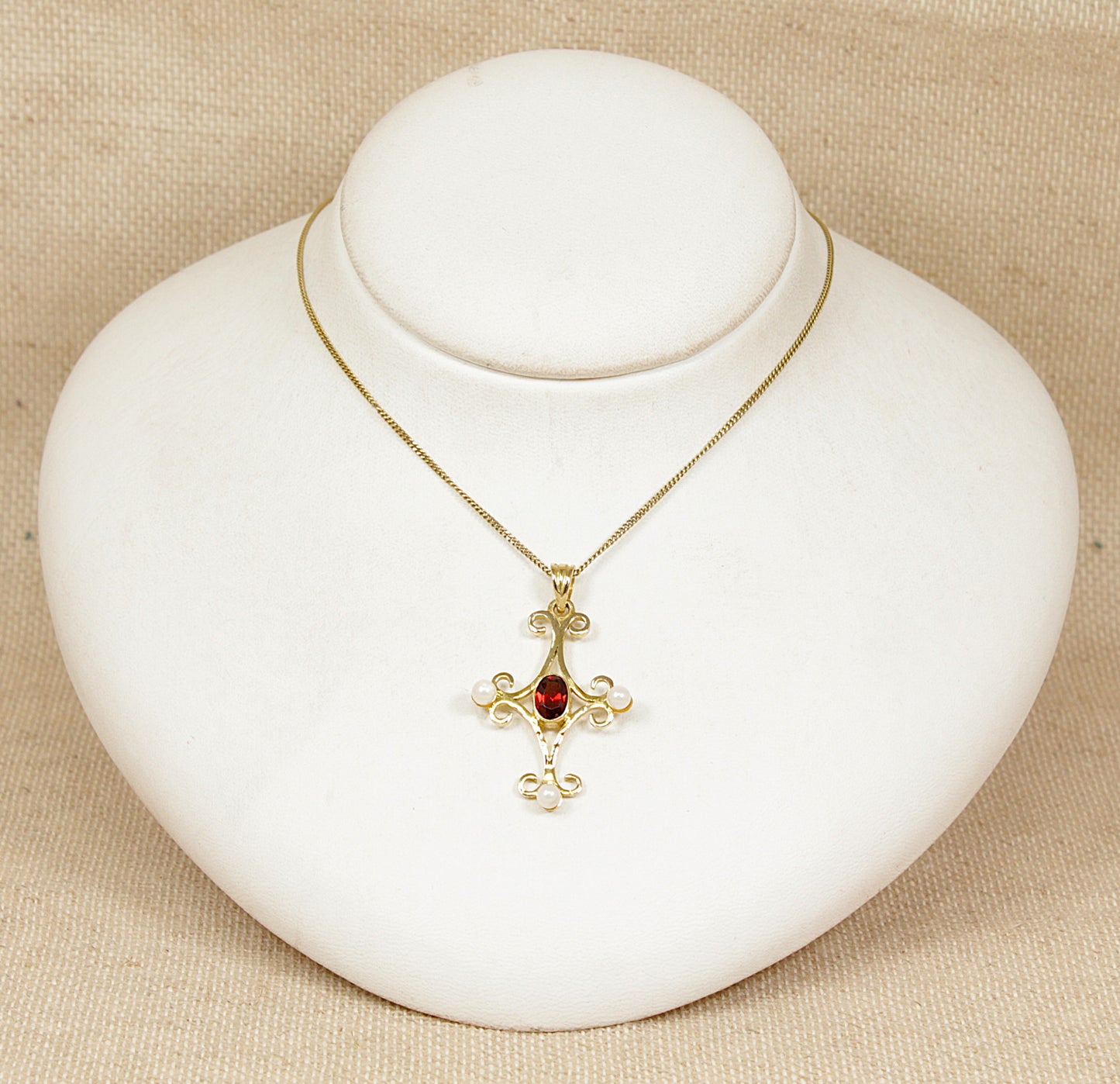 Silver gold plated  necklace with a cross pendant featuring a red garnet and pearl gemstones on a white stand.