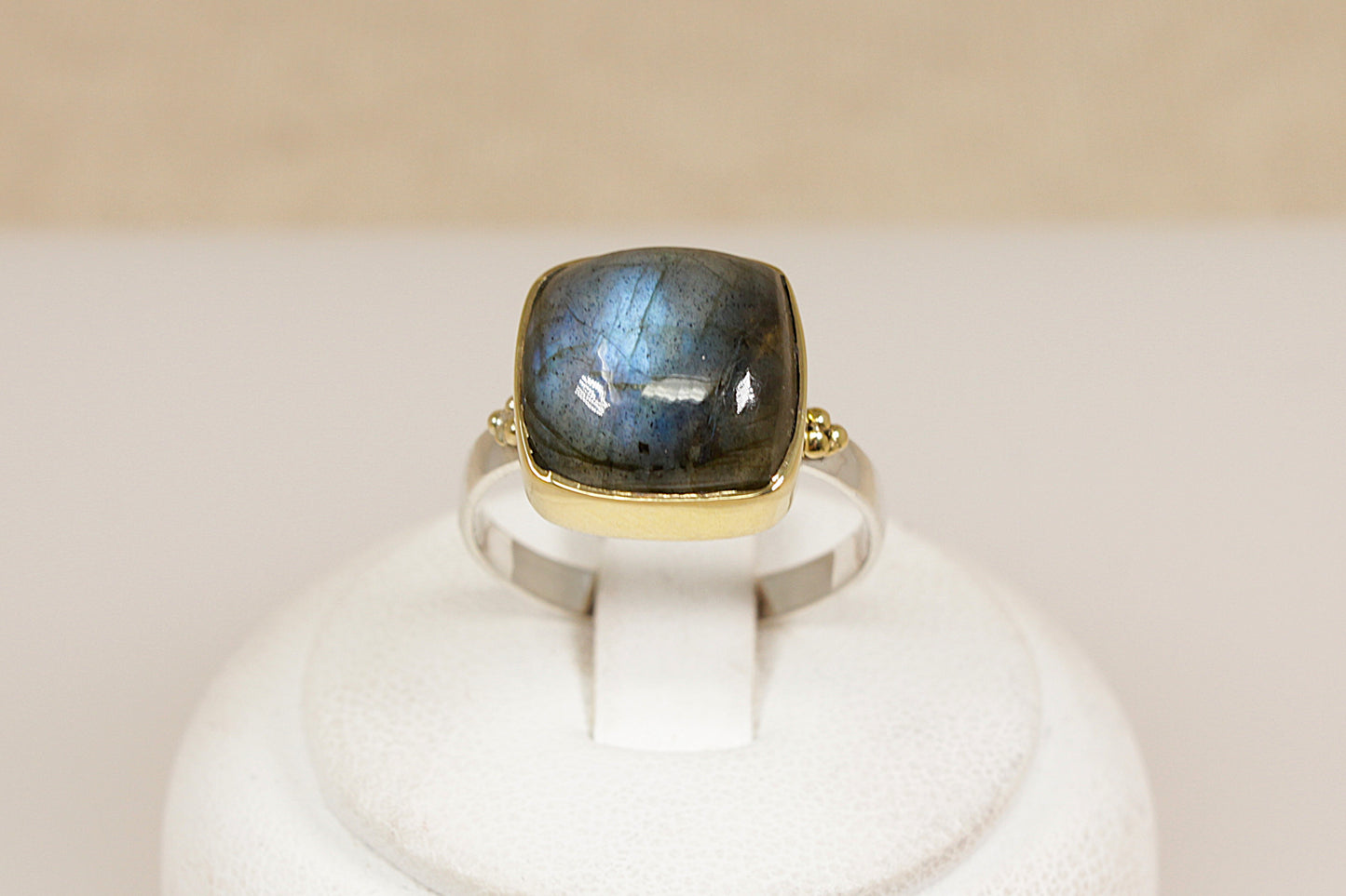 Ring with a labradorite gemstone and gold band on a white stand against a beige background