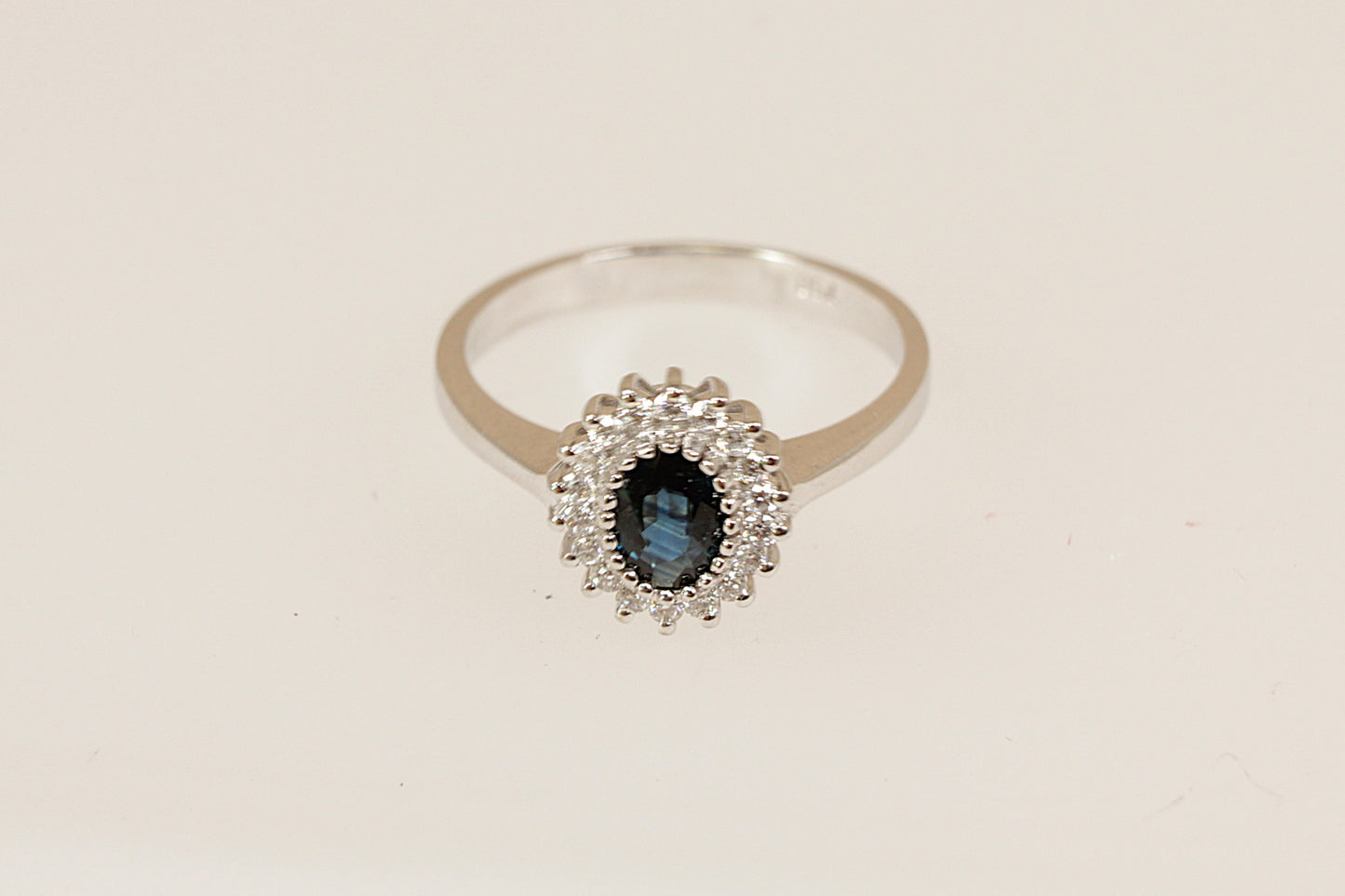 18ct white gold Diana style ring with a blue sapphire gemstone surrounded by a halo of diamonds on a beige background