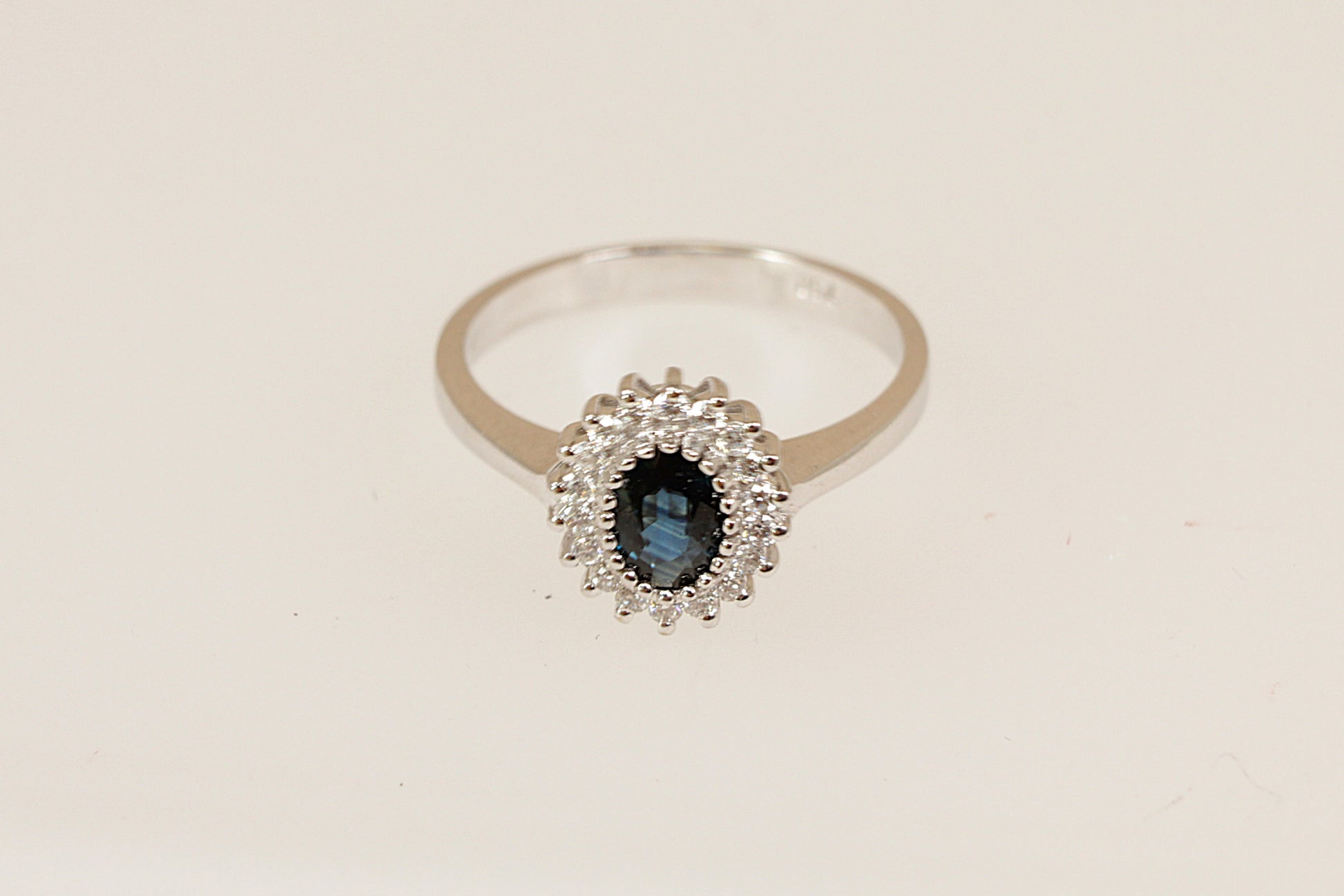 18ct white gold Diana style ring with a blue sapphire gemstone surrounded by a halo of diamonds on a beige background