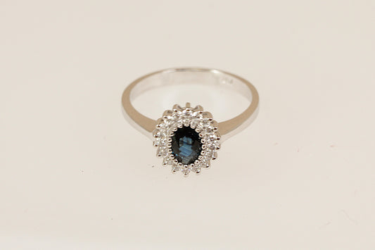 18ct white gold Diana style ring with a blue sapphire gemstone surrounded by a halo of diamonds on a beige background