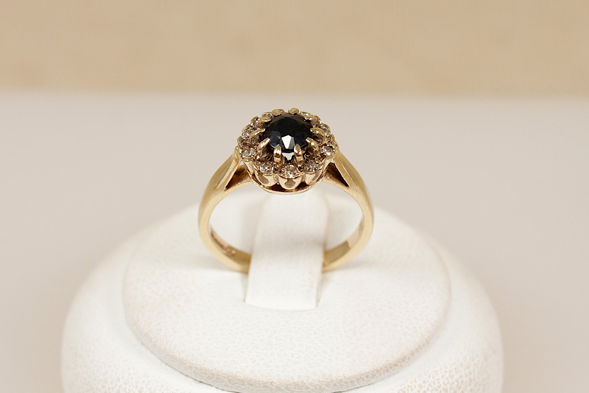 Gold ring with a central blue sapphire  gemstone surrounded by a halo of diamonds on a white stand against a beige background