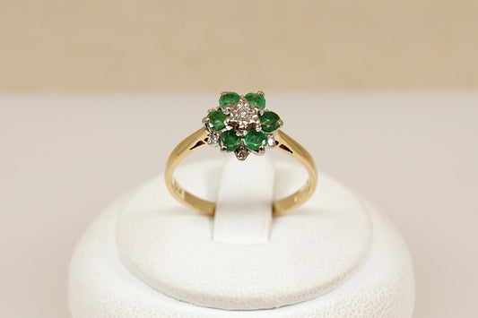 Gold ring with emerald and diamond cluster on a white stand against a beige background