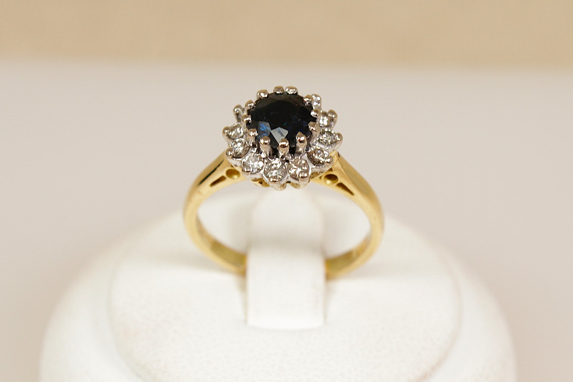 Gold ring with a central blue sapphire  gemstone and diamond-studded halo on a white stand.
