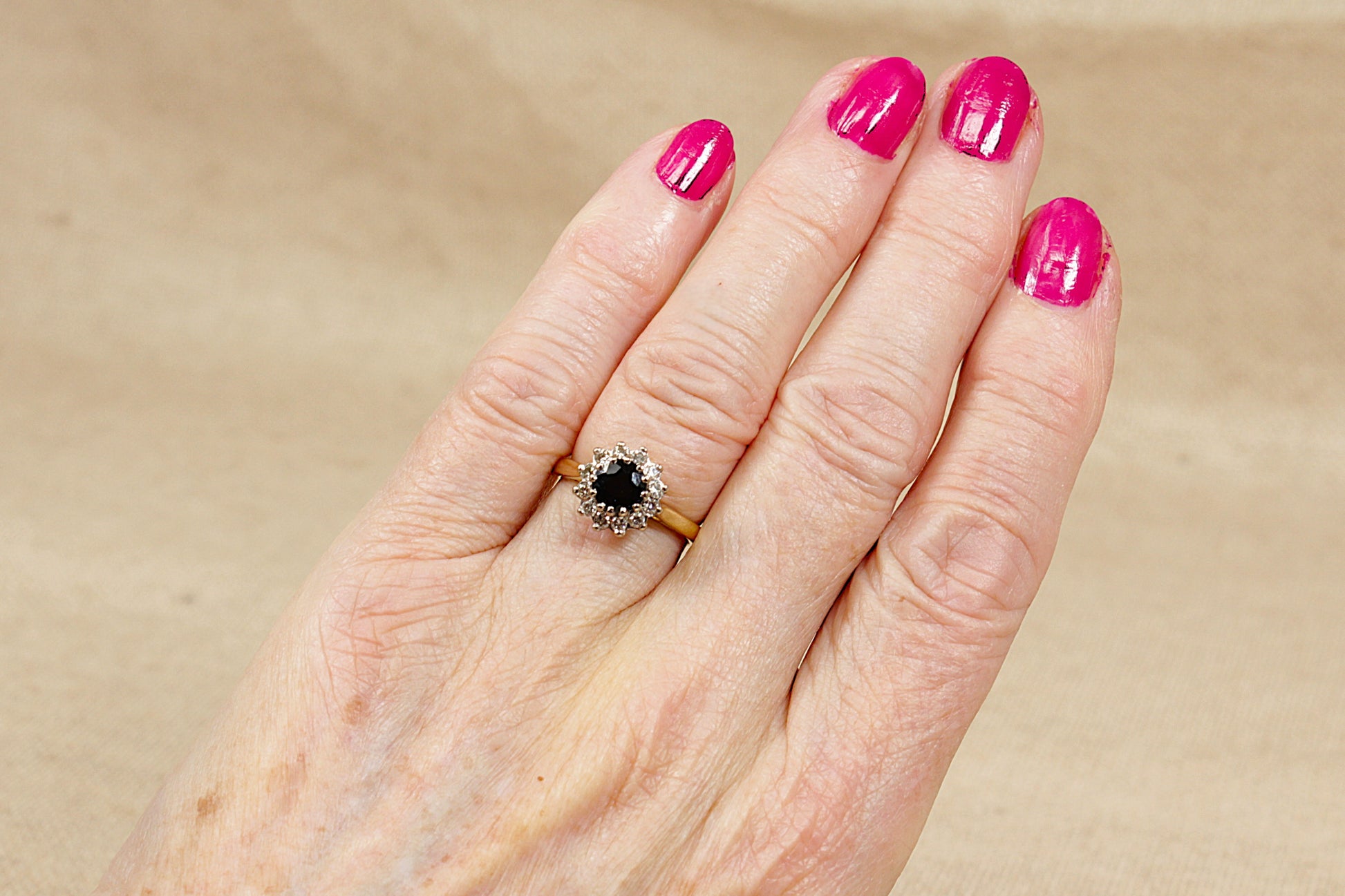 Hand with pink nail polish wearing a sapphire and diamond gold ring on a beige background
