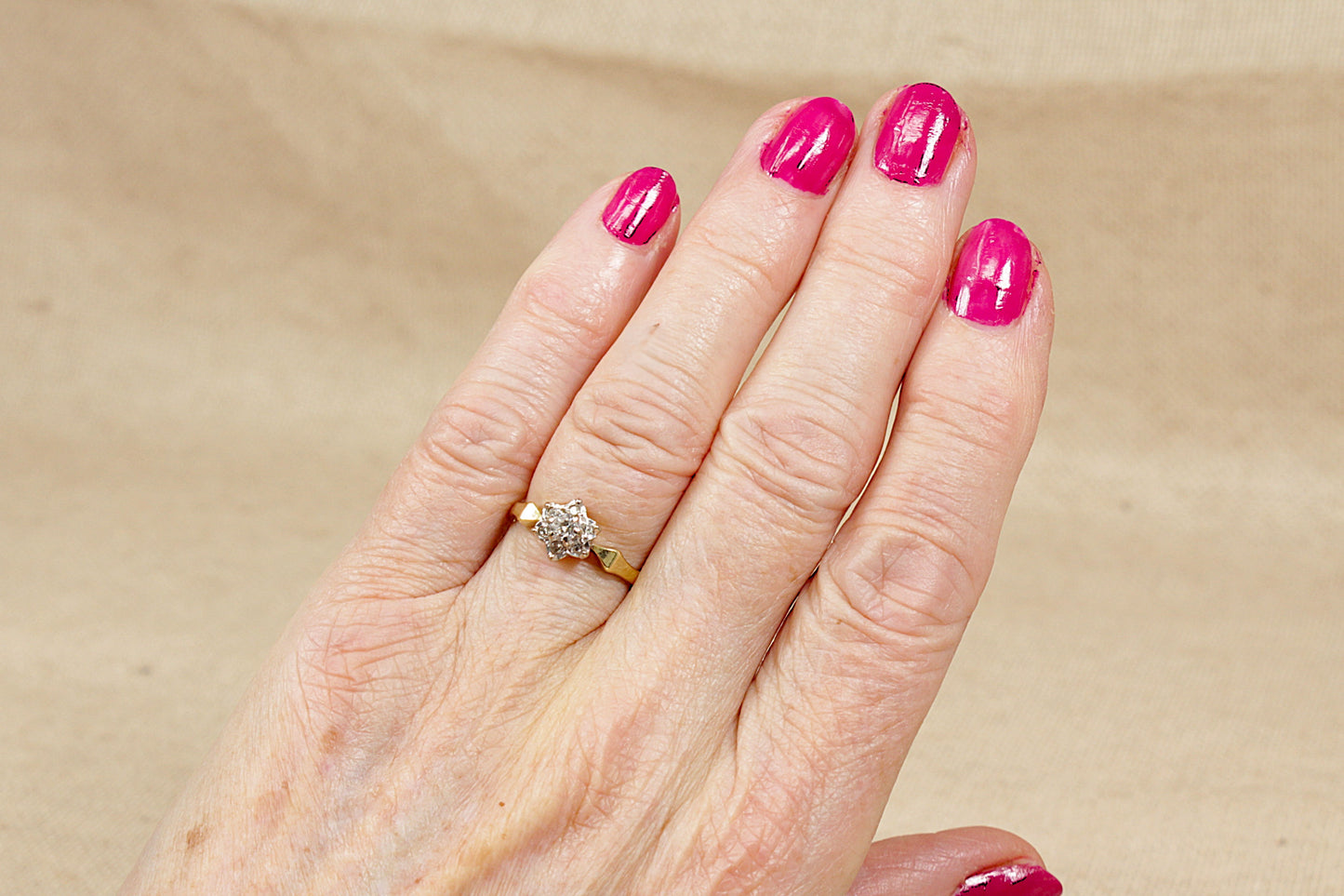 Hand with pink nail polish wearing a gold diamond cluster ring on a beige background