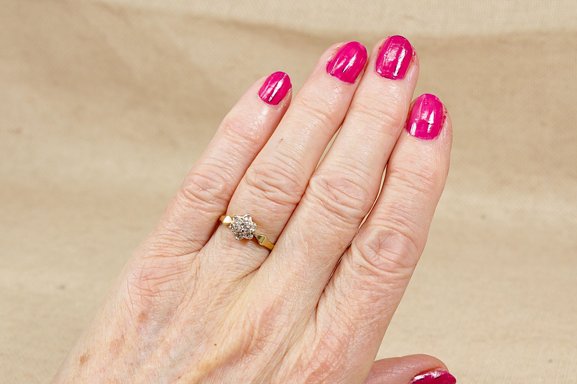 Hand with pink nail polish wearing a gold diamond cluster ring on a beige background