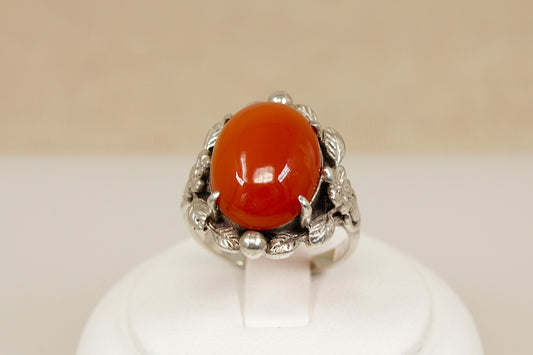 Silver arts and crafts ring with a large red carnelian gemstone on a white stand against a beige background