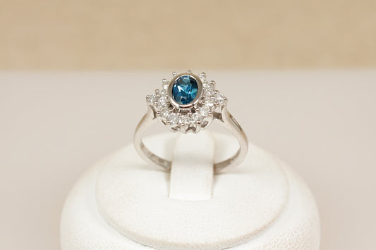 18ct white gold  ring with a blue topaz gemstone surrounded by diamonds on a white stand against a beige background