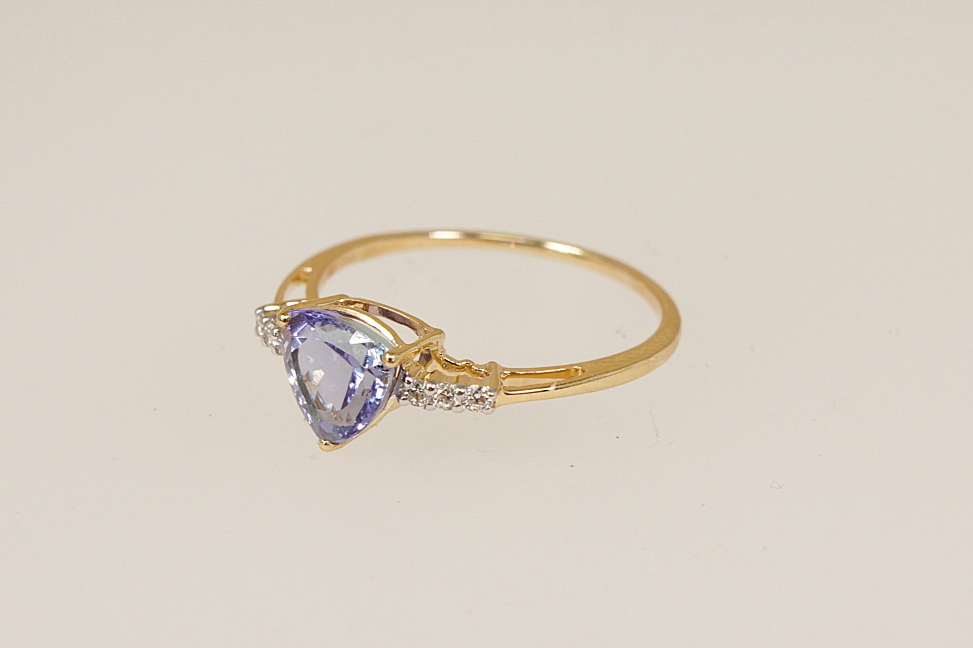 Gold ring with a trillion cut tanzanite  gemstone with diamond accents on each shoulder on a beige background