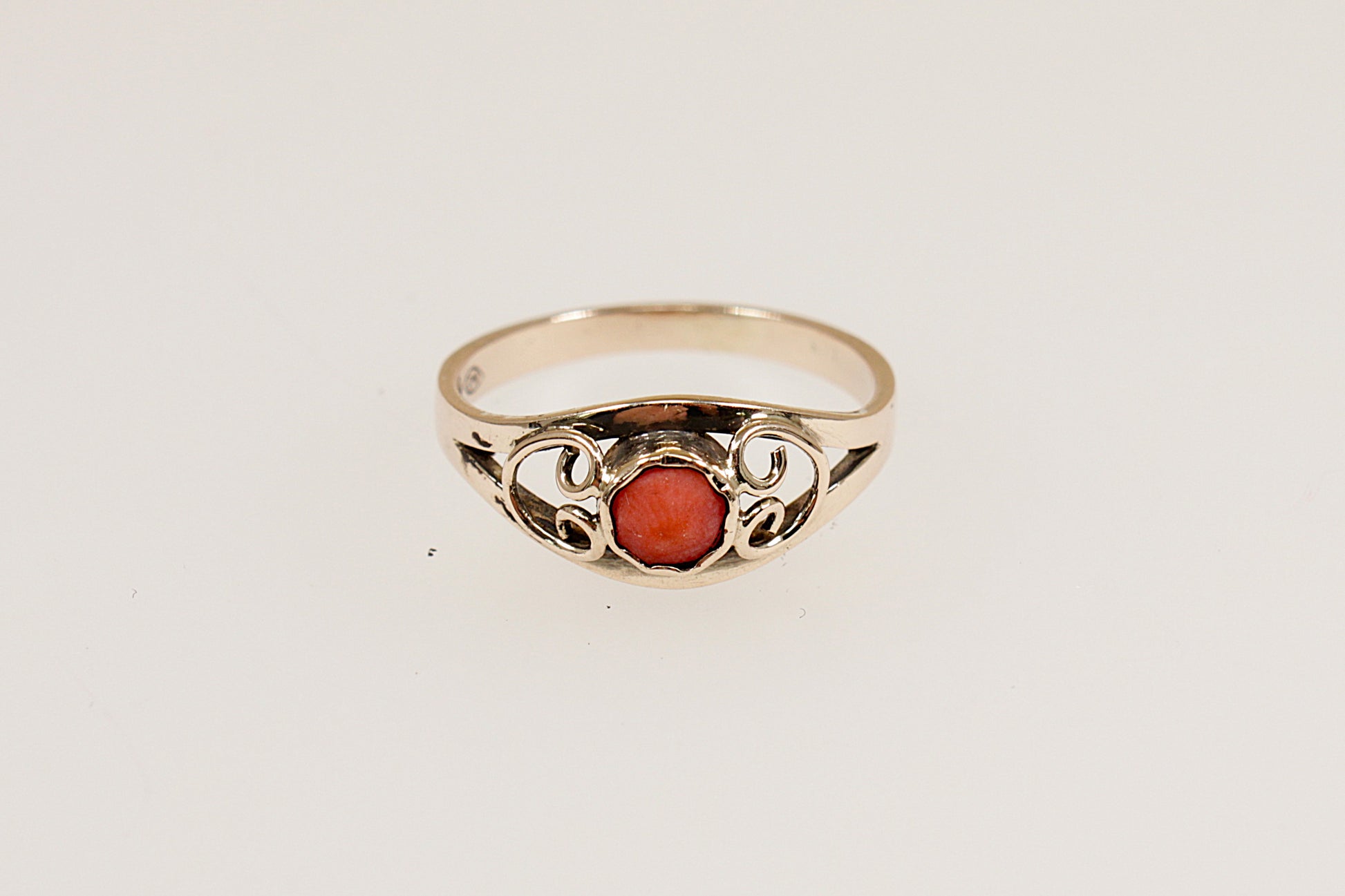 A gold ring with a round coral centerpiece set in a scalloped bezel, with gold heart designs on each shoulder, displayed against a white background.