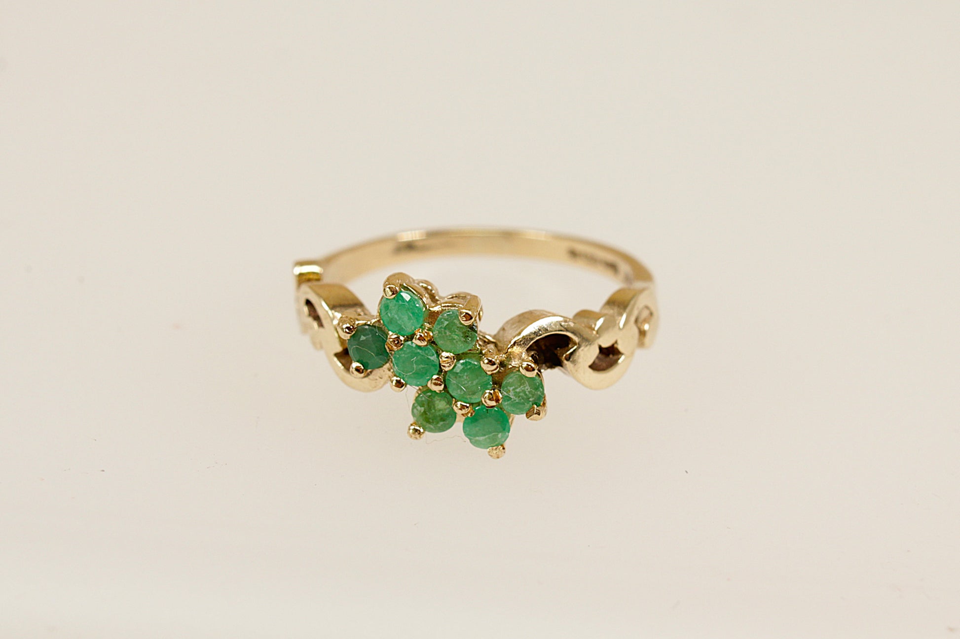 A charming vintage cluster ring crafted in warm 9ct yellow gold, featuring eight round-cut emeralds arranged in a delicate floral design. Each emerald is securely claw-set, creating a vibrant cluster that radiates natural green beauty. The ring's decorative shoulders are adorned with elegant scrolling goldwork, adding a touch of ornate detail and vintage character. Inside the band are full London hallmarks, with the date letter for 1979 and the maker’s mark ‘R & W’. UK finger size N.