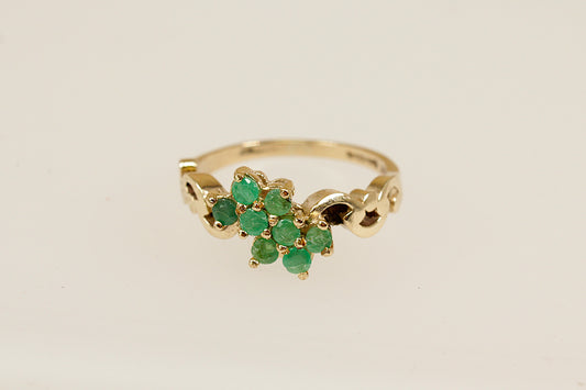 A charming vintage cluster ring crafted in warm 9ct yellow gold, featuring eight round-cut emeralds arranged in a delicate floral design. Each emerald is securely claw-set, creating a vibrant cluster that radiates natural green beauty. The ring's decorative shoulders are adorned with elegant scrolling goldwork, adding a touch of ornate detail and vintage character. Inside the band are full London hallmarks, with the date letter for 1979 and the maker’s mark ‘R & W’. UK finger size N.