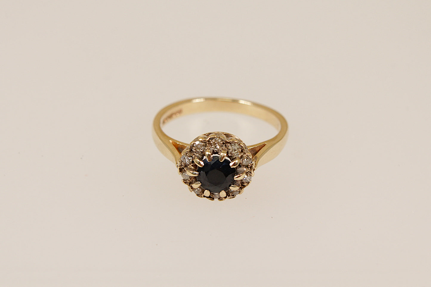 Gold ring with a central blue sapphire  gemstone surrounded by a halo of diamonds on a white stand against a beige background