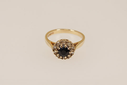 Gold ring with a central blue sapphire  gemstone surrounded by a halo of diamonds on a white stand against a beige background