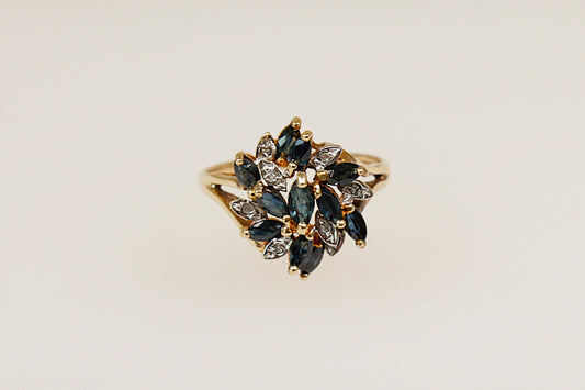 This 9ct Gold ring is a breathtaking and artistic statement piece! This large Sapphire and Diamond flower design ring features 11 Marquise-cut blue Sapphires in claw settings and 8 round diamond accents, Birmingham hallmarks for year 1994 and stamped DIA. UK finger size O. 