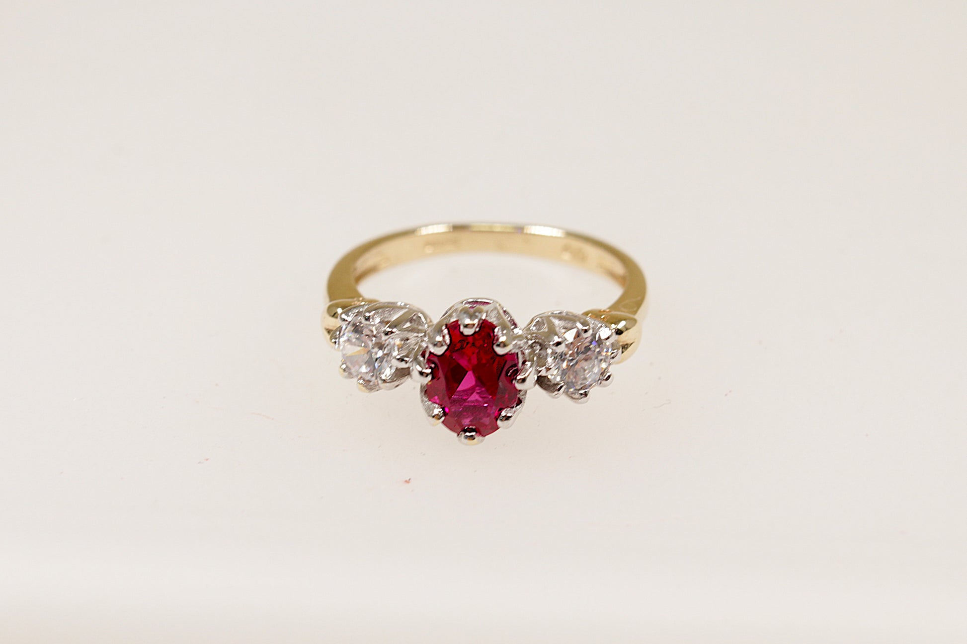 A 9ct gold trilogy ring with an oval cut ruby centerpiece and two round cut crystal gemstones on either side, set in a crown-inspired setting.
