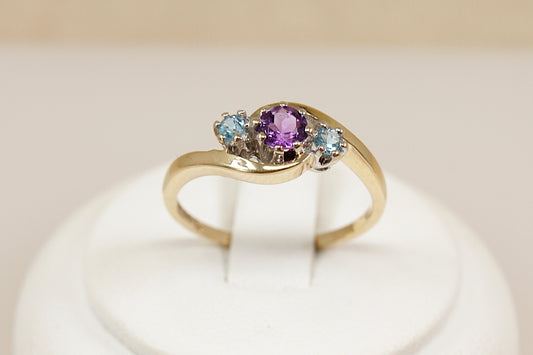 A delightful and feminine 9ct gold trilogy ring, featuring a round claw-set Amethyst at the centre, flanked by two sparkling aquamarines on either side. The gemstones are elegantly arranged in a twist setting, giving the design a graceful flow and a touch of vintage romance, The ring is hallmarked in London for 1994, confirming its gold purity and vintage authenticity, UK Finger size: O.