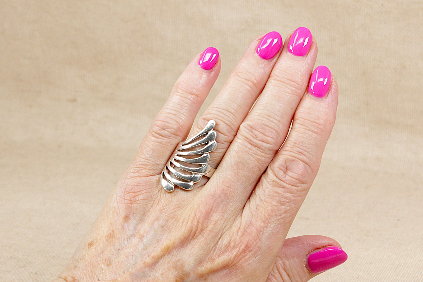 Hand wearing an abstract silver statement ring with pink nail polish on a beige background