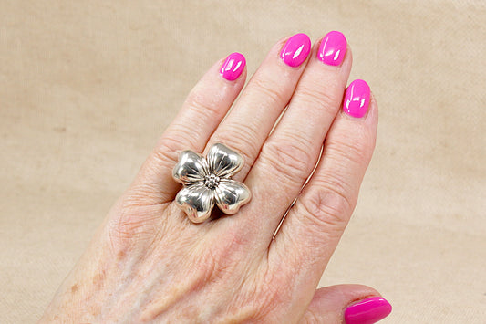 Silver statement ring in the form of a four leaf clover flower on a ladies finger with pink nail varnish with a beige background