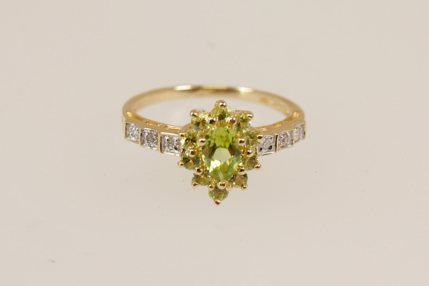 A vibrant and eye-catching 9ct gold ring, featuring a beautifully cut pear-shaped peridot at its centre. Surrounding the main stone is a sparkling halo of 10 smaller peridots, all claw set in an elegant basket setting that enhances the ring’s depth and brilliance. The stepped shoulders are adorned with delicate diamond accents, adding a refined sparkle and completing the luxurious design. Inside the band, the ring is clearly stamped 375 and 9K, confirming its gold purity. UK Finger size: N ½.