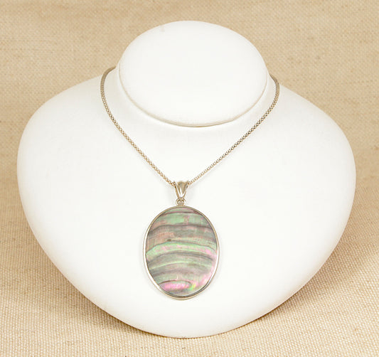 A beautifully eye-catching large sterling silver pendant featuring an iridescent oval abalone shell, bezel-set to allow visibility of both the front and back. The abalone shimmers with an array of blues, greens, and purples, catching the light with every movement and making each piece naturally unique. The pendant sits on a silver 18 inch chain.