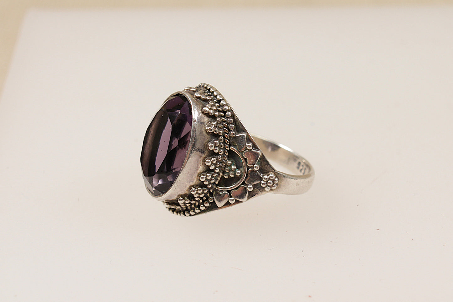 A silver ring with an amethyst coloured central stone and intricate silver patterns around it, displayed against a white background.