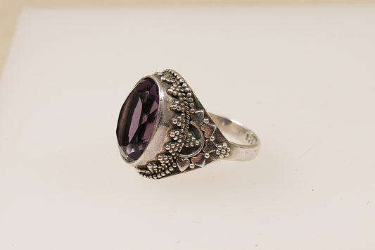 A silver ring with an amethyst coloured central stone and intricate silver patterns around it, displayed against a white background.