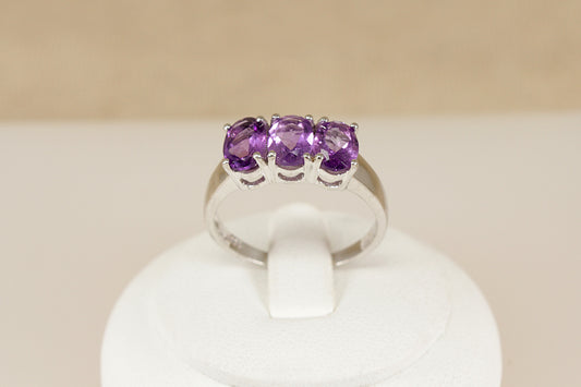 Silver ring with three purple amethyst   gemstones on a white stand against a beige background