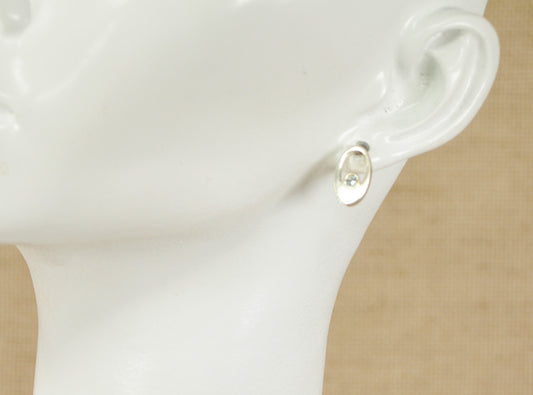 A pair of silver stud earrings with blue topaz displayed on a mannequin ear.
