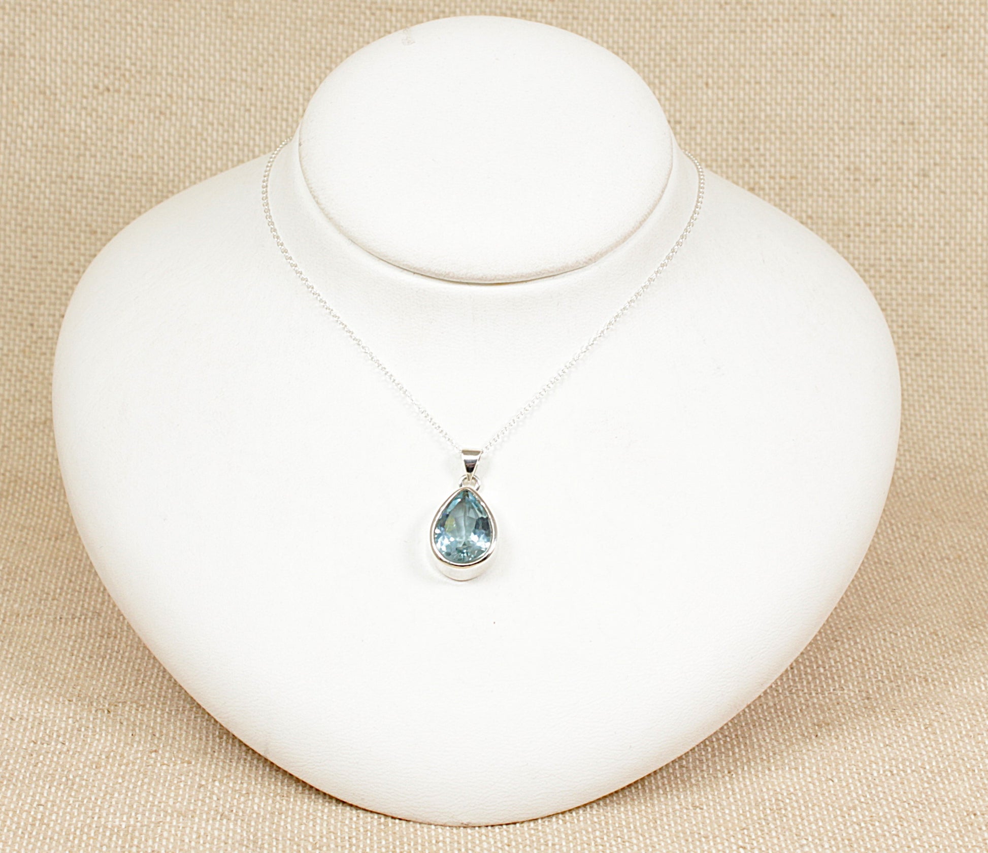A silver teardrop pendant necklace with a blue topaz gemstone displayed on a white bust.