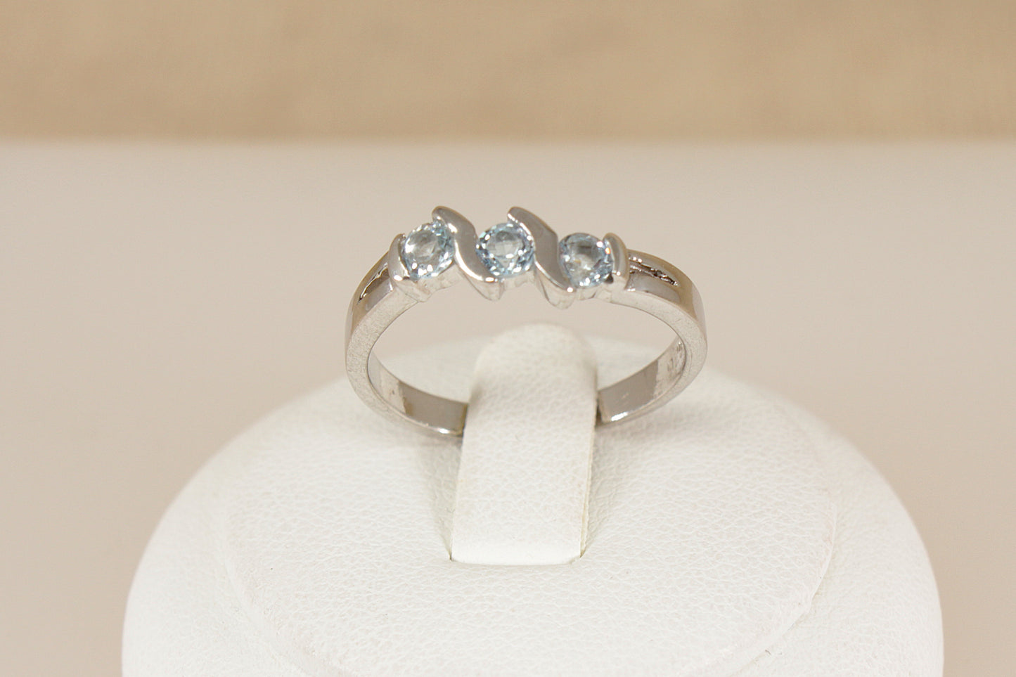 Silver ring with three blue topaz gemstones on a white stand against a beige background