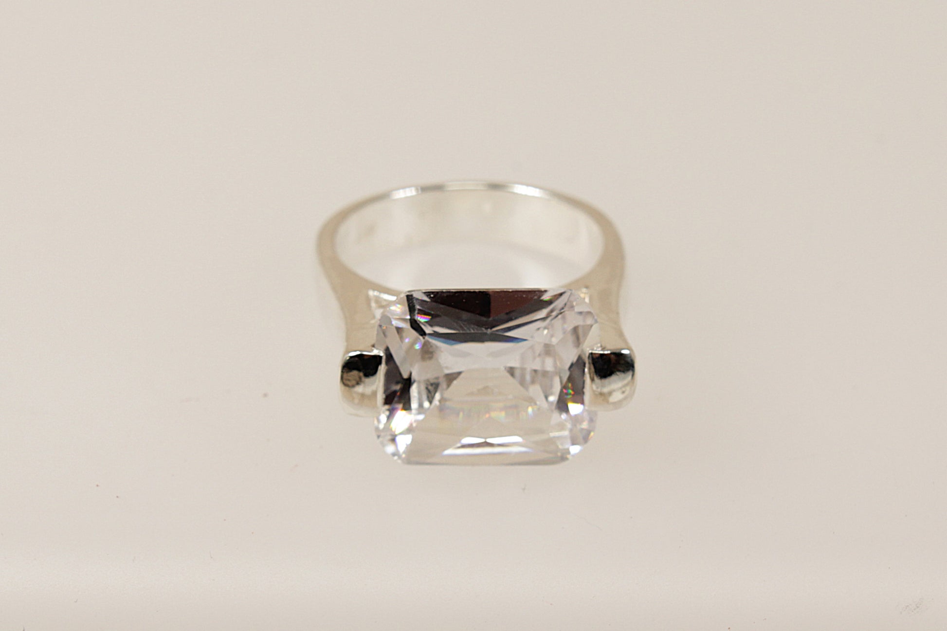 This silver CZ ring is a bold and striking piece, characterised by its substantial design and unique architectural setting.
The main feature is the large emerald cut Cubic Zirconia gemstone set in a cradle style mount, the stone is secured with 2 chunky claws that extend from the shoulders of the ring. The combination of the large CZ, heavy silver band, and innovative design makes this ring perfect for those who appreciate modern eye-catching statement jewellery. 

It is in excellent condition and inside t