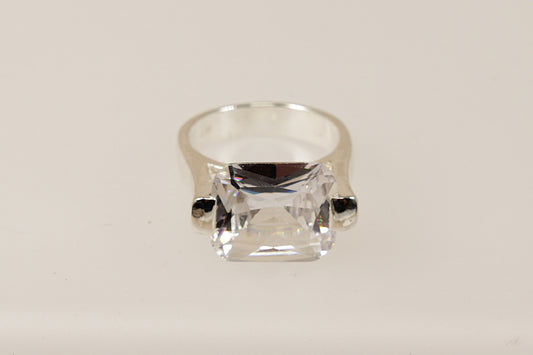 This silver CZ ring is a bold and striking piece, characterised by its substantial design and unique architectural setting.
The main feature is the large emerald cut Cubic Zirconia gemstone set in a cradle style mount, the stone is secured with 2 chunky claws that extend from the shoulders of the ring. The combination of the large CZ, heavy silver band, and innovative design makes this ring perfect for those who appreciate modern eye-catching statement jewellery. 

It is in excellent condition and inside t