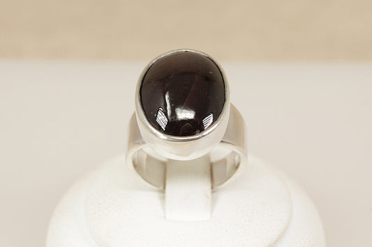 A striking and beautifully crafted sterling silver statement ring, showcasing a large cabochon-cut garnet with a rich, deep red hue. The garnet is set in a deep-sided bezel, giving the piece a bold, sculptural presence while highlighting the smooth, glossy surface of the gemstone. UK finger size O.