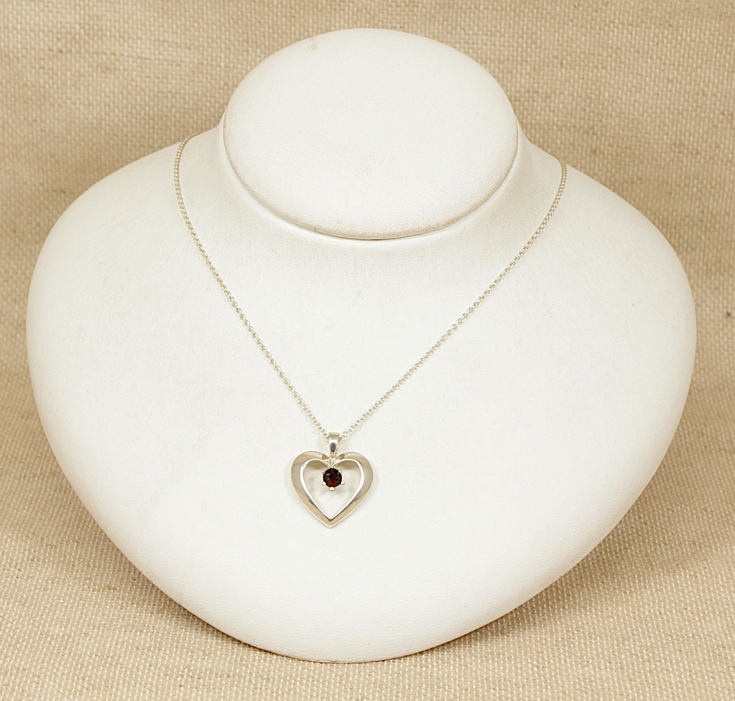 Silver necklace with a heart-shaped pendant with garnet gemstone on a white stand against a beige background