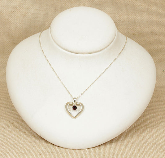 Silver necklace with a heart-shaped pendant with garnet gemstone on a white stand against a beige background
