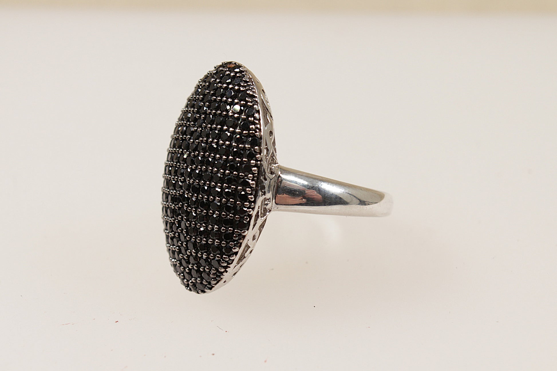This Silver ring is a beautiful and bold jewellery piece; it is a large oval shape with a domed basket that creates an elegant elevated profile. The surface of the of the ring is pave` set with an array of glittery Garnet gemstones giving it a luxurious and radiant sparkle. The underside or gallery of the ring features intricate filigree work, adding depth and elegance to the overall look. UK finger size O.