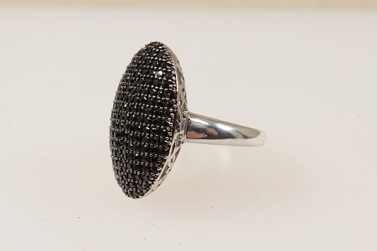 This Silver ring is a beautiful and bold jewellery piece; it is a large oval shape with a domed basket that creates an elegant elevated profile. The surface of the of the ring is pave` set with an array of glittery Garnet gemstones giving it a luxurious and radiant sparkle. The underside or gallery of the ring features intricate filigree work, adding depth and elegance to the overall look. UK finger size O.