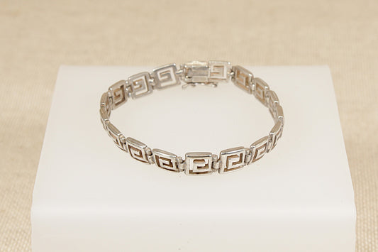 A silver bracelet with a Greek Key design, featuring square articulated links, displayed on a black background.