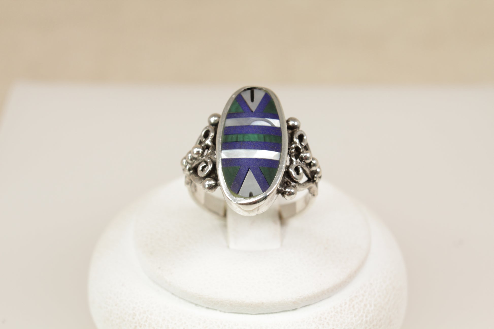 A striking and artistic sterling silver ring, featuring a large oval bezel setting inlaid with a geometric mosaic of Malachite, Mother of Pearl, and Lapis Lazuli. The smooth, polished surface creates a vibrant interplay of colour and texture—from the rich green of the Malachite, to the soft iridescence of mother of pearl, and the deep royal blue of Lapis Lazuli, uk finger size M.