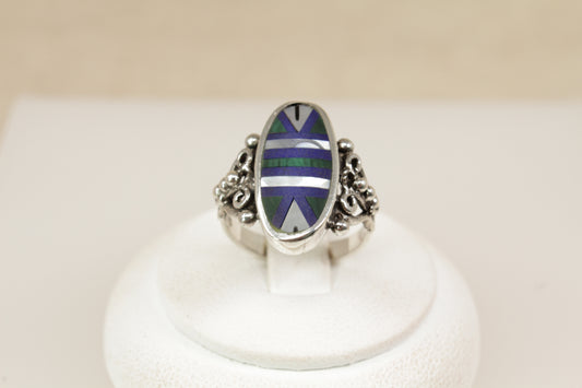 A striking and artistic sterling silver ring, featuring a large oval bezel setting inlaid with a geometric mosaic of Malachite, Mother of Pearl, and Lapis Lazuli. The smooth, polished surface creates a vibrant interplay of colour and texture—from the rich green of the Malachite, to the soft iridescence of mother of pearl, and the deep royal blue of Lapis Lazuli, uk finger size M.