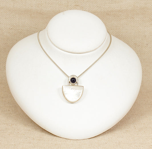 A striking and substantial sterling silver pendant showcasing a sleek, modernist design. The piece features a luminous semicircle of Mother of Pearl, bezel-set to highlight its natural iridescence. Above it rests a round cabochon of deep violet Iolite, adding a touch of rich colour and contrast to the composition. 
16 inches long.