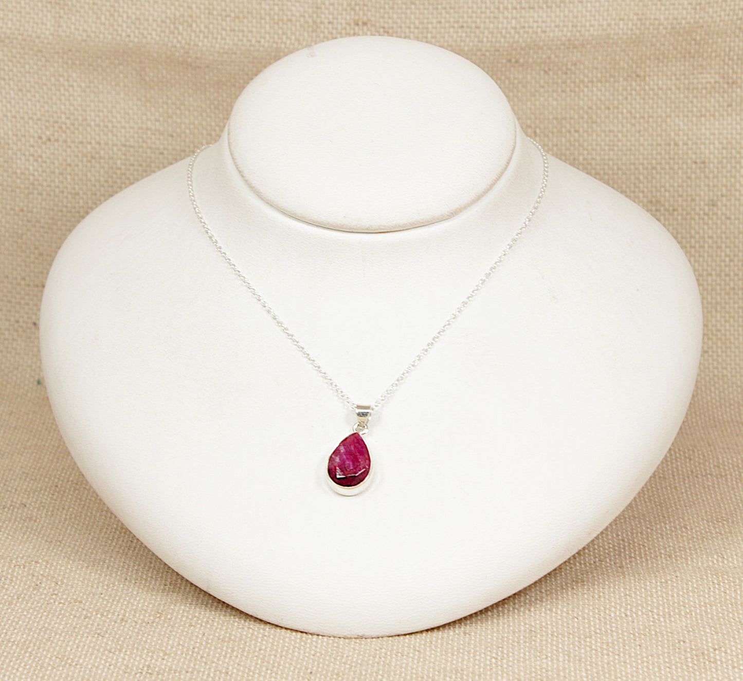 Silver necklace with a teardrop-shaped ruby pendant on a white stand against a beige background